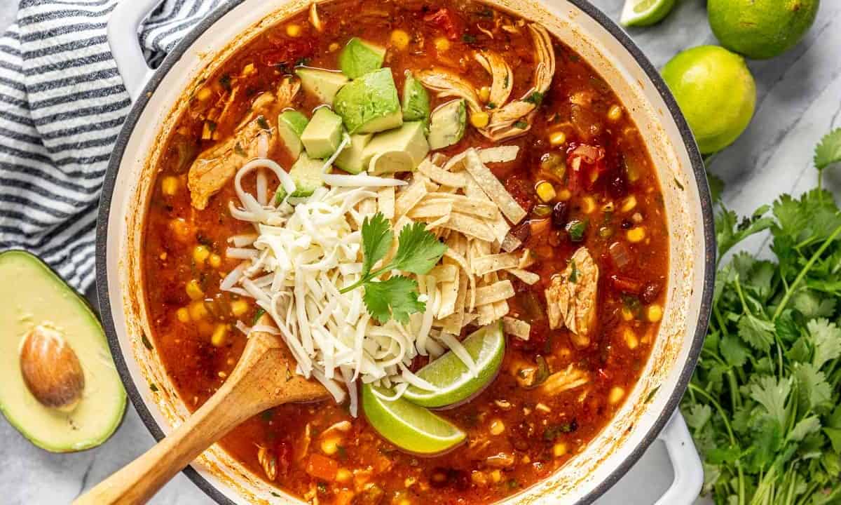 Bird's eye view of chicken tortilla soup topped with cheese, avocado, tortilla strips, lime, and cilantro, served out of a large dutch oven pot with a wooden spoon.