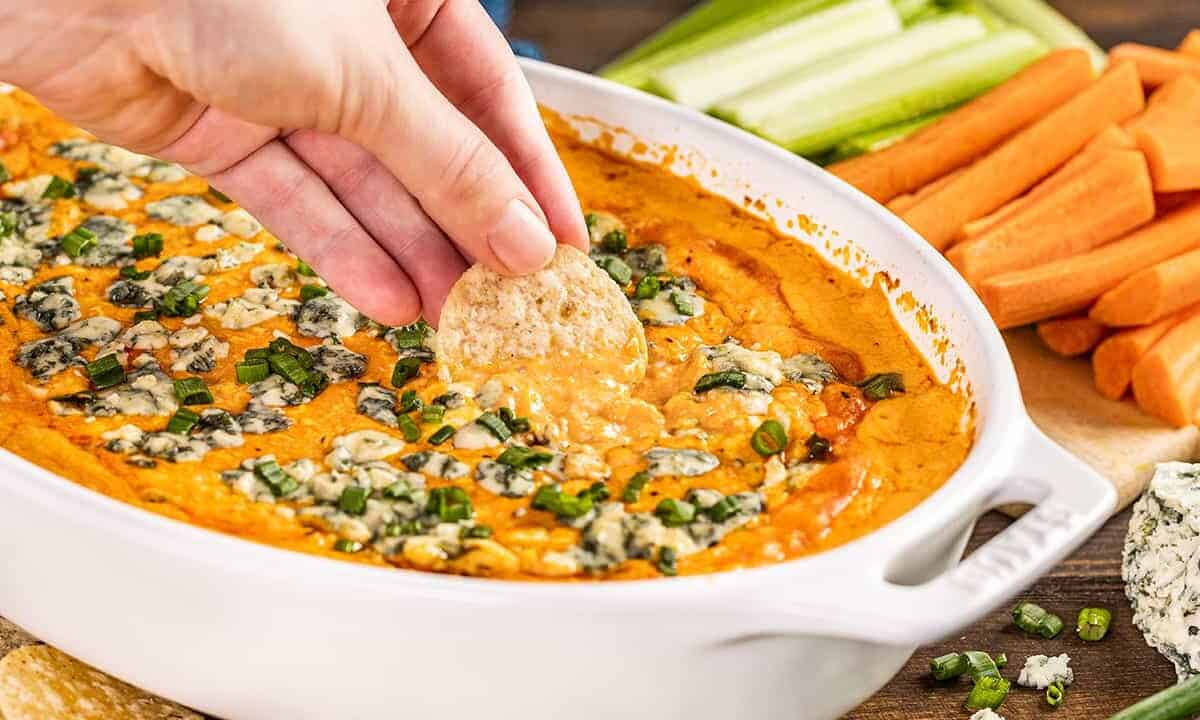 a hand dipping a cracker into buffalo chicken dip in a baking dish.