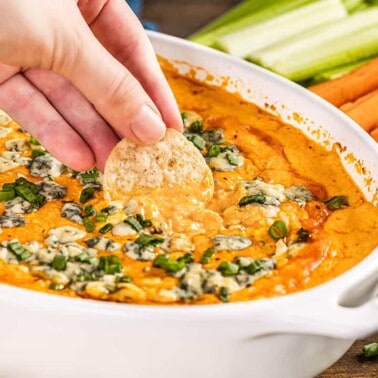 a hand dipping a cracker into buffalo chicken dip in a baking dish.