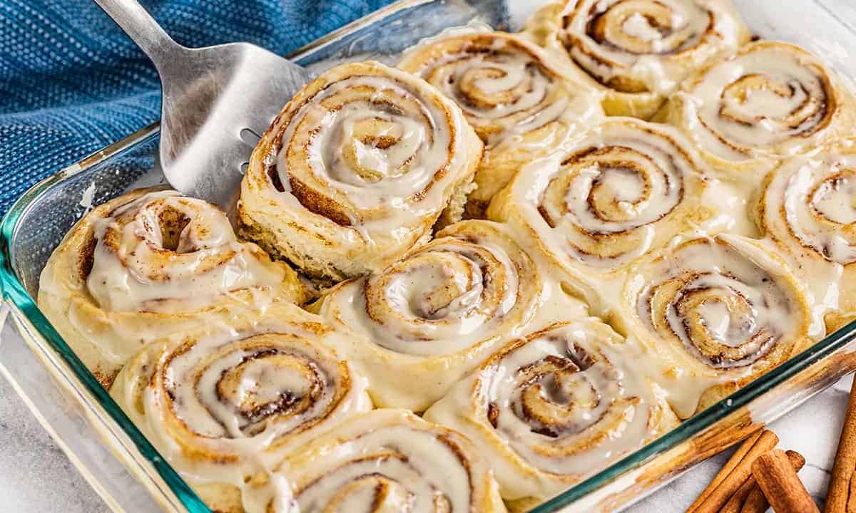 A spatula picking up a cinnamon roll from a glass baking pan.