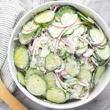 Bird's eye view of creamy cucumber salad in a white bowl with a wooden spoon next to it.