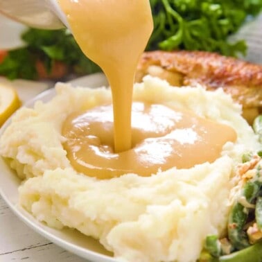 Gravy poured from a gravy boat onto a plate of mashed potatoes next to roasted turkey and green beans.