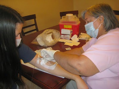 A nurse working on a PICC line on Rachel's arm while sitting at a kitchen table covered with medical supplies.