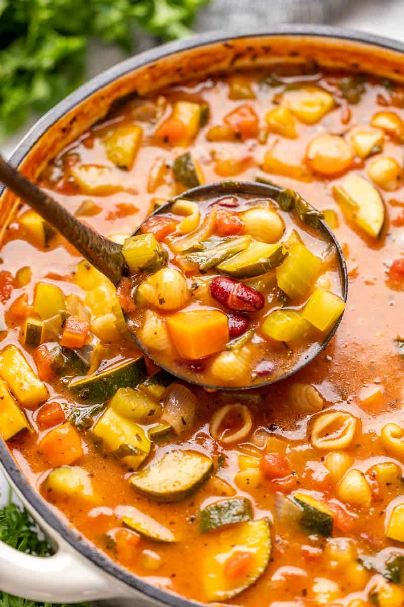 Overhead view of a white pot full of minestrone soup with a ladle full of soup being scooped out.